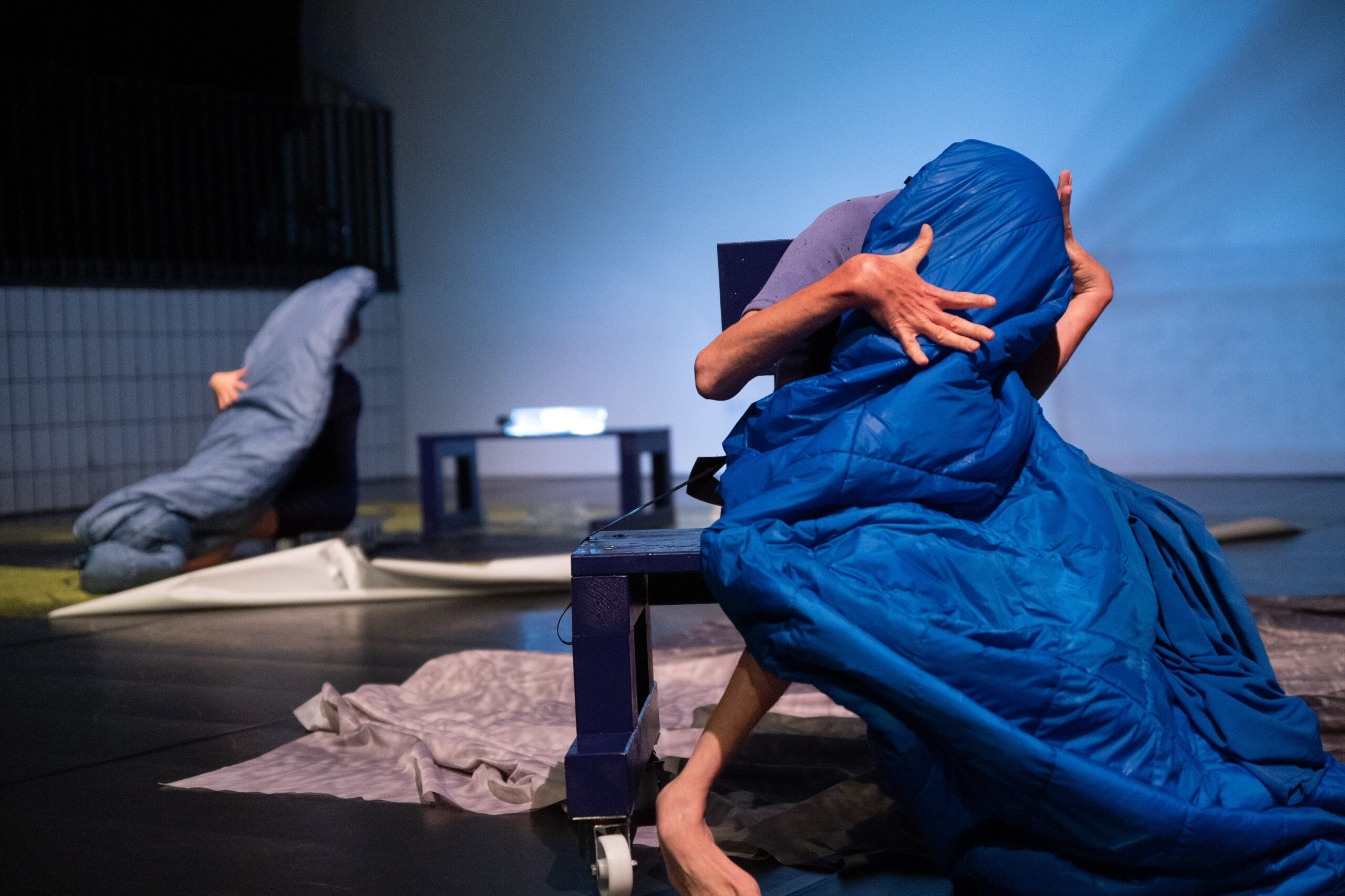 Image description: A performer sits on a blue bench, their face and most of their body covered by a blue sleeping bag. Their hands and one foot, the only parts of them that are visible, are tense, extended wide and folding in towards their shrouded body. Behind them, out of focus, a second dancer sits behind a sleeping bag in a similar position. Around them both, the ground is scattered with large grey sheets.
