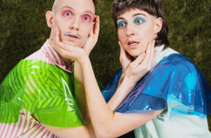 Image description: Two performers with expressions of frozen panic crouch towards each other. Dressed in bold colours with bright eyeshadow and drawn-on moustahces, each one cups the other's face between their hands.