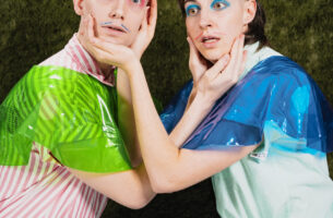 Image description: Two performers with expressions of frozen panic crouch towards each other. Dressed in bold colours with bright eyeshadow and drawn-on moustahces, each one cups the other's face between their hands.