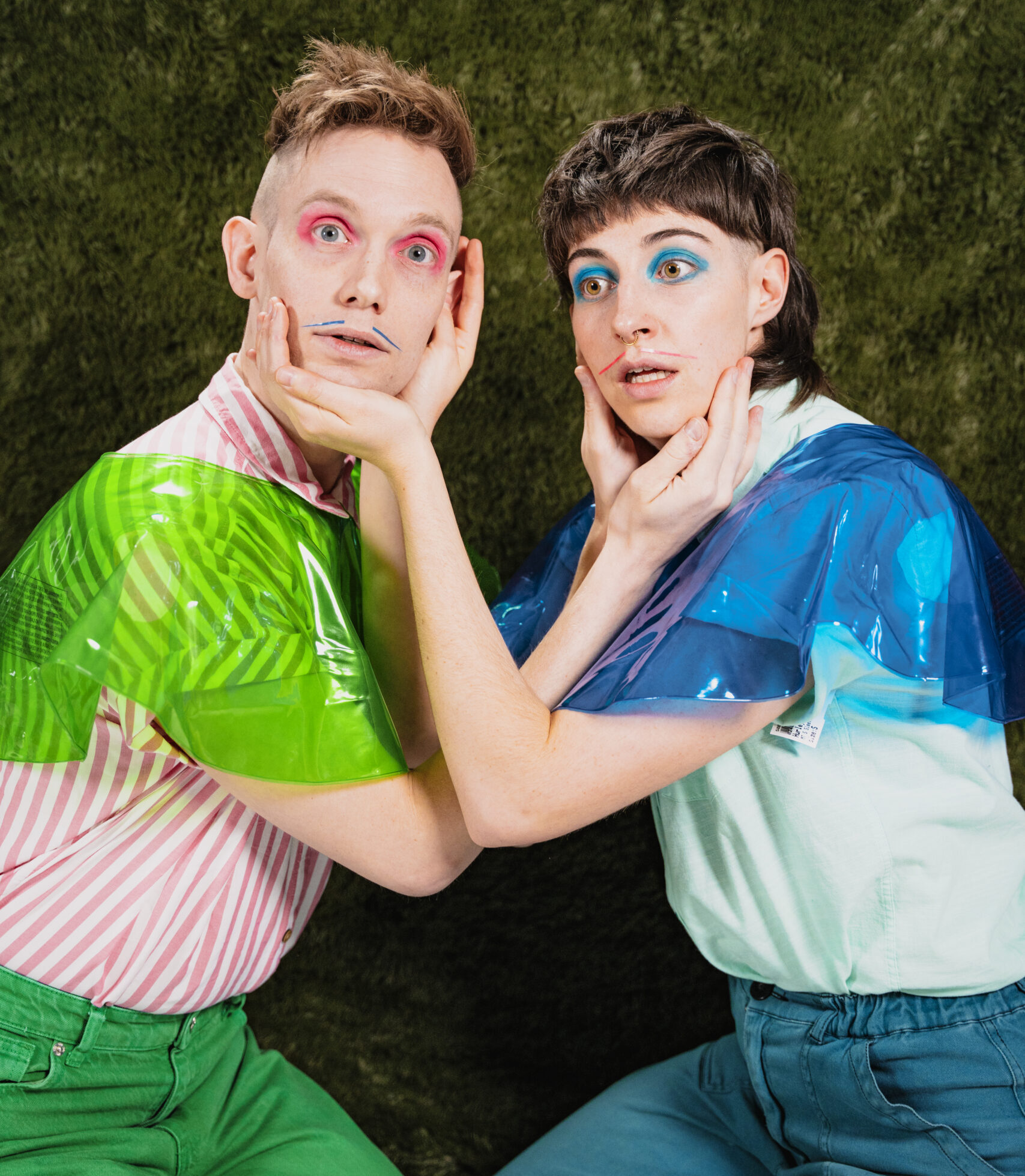 Image description: Two performers with expressions of frozen panic crouch towards each other. Dressed in bold colours with bright eyeshadow and drawn-on moustahces, each one cups the other's face between their hands.