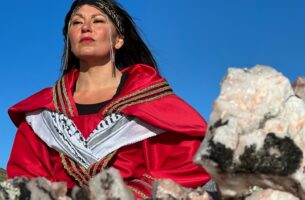 Image description: Outdoors, an Indigenous woman looks proudly into the distance. She wears an embroidered headband, an ornate red coat, and a Palestinian keffiyeh tucked neatly around her shoulders under the oppulent folds of her coat's collar. Behind her is a cloudless blue sky.