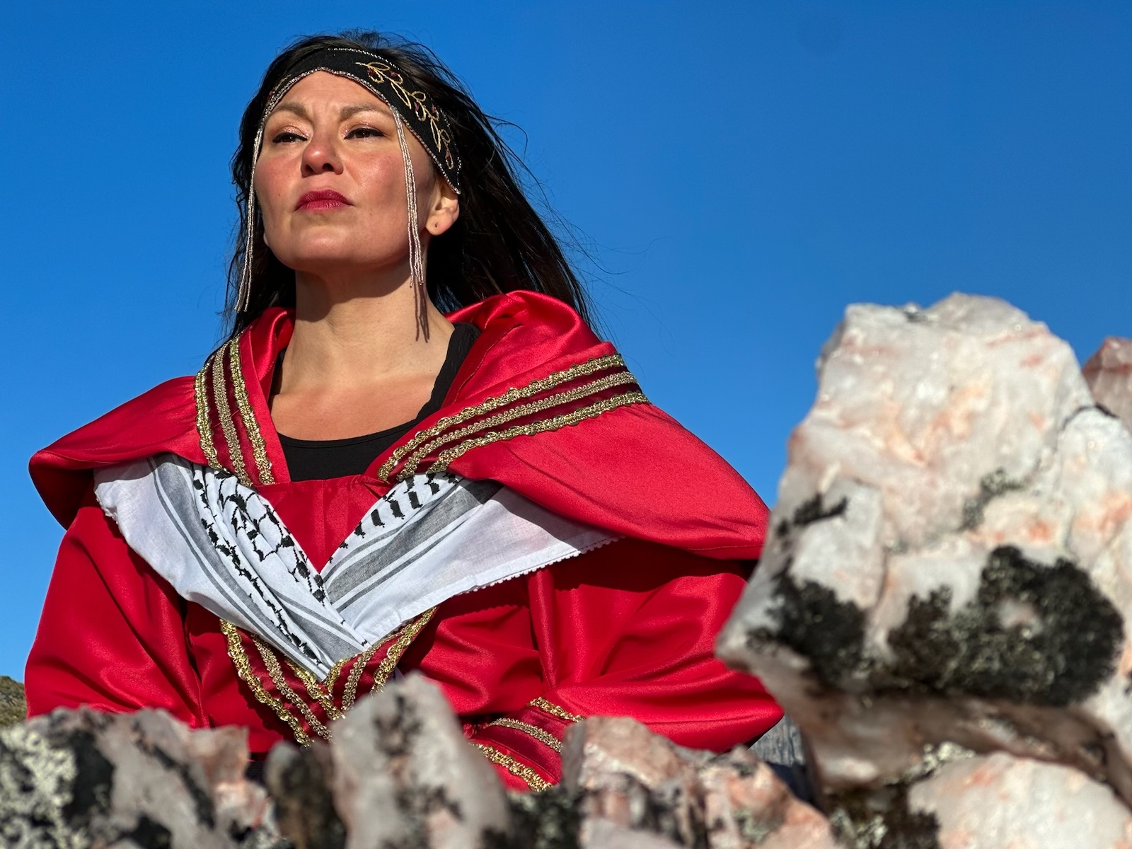 Image description: Outdoors, an Indigenous woman looks proudly into the distance. She wears an embroidered headband, an ornate red coat, and a Palestinian keffiyeh tucked neatly around her shoulders under the oppulent folds of her coat's collar. Behind her is a cloudless blue sky.