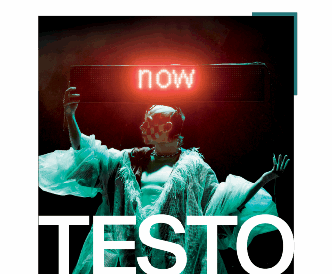 TESTO: A trans-masculine drag artist stands on a dark stage, holding an LED-sign where the word "now" glows red. Their bald head is turned in profile, with a checkerboard smile painted across their face. They wear a billowing white coat made of luxuriously gauzy material, which hangs open, exposing a flat, muscular chest.