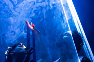 Image description: Two performers stand close to each other, eyes looking upward pensively between beams of blue light, filled with swirling haze. One reaches her fingers into the light, like dipping into another realm.