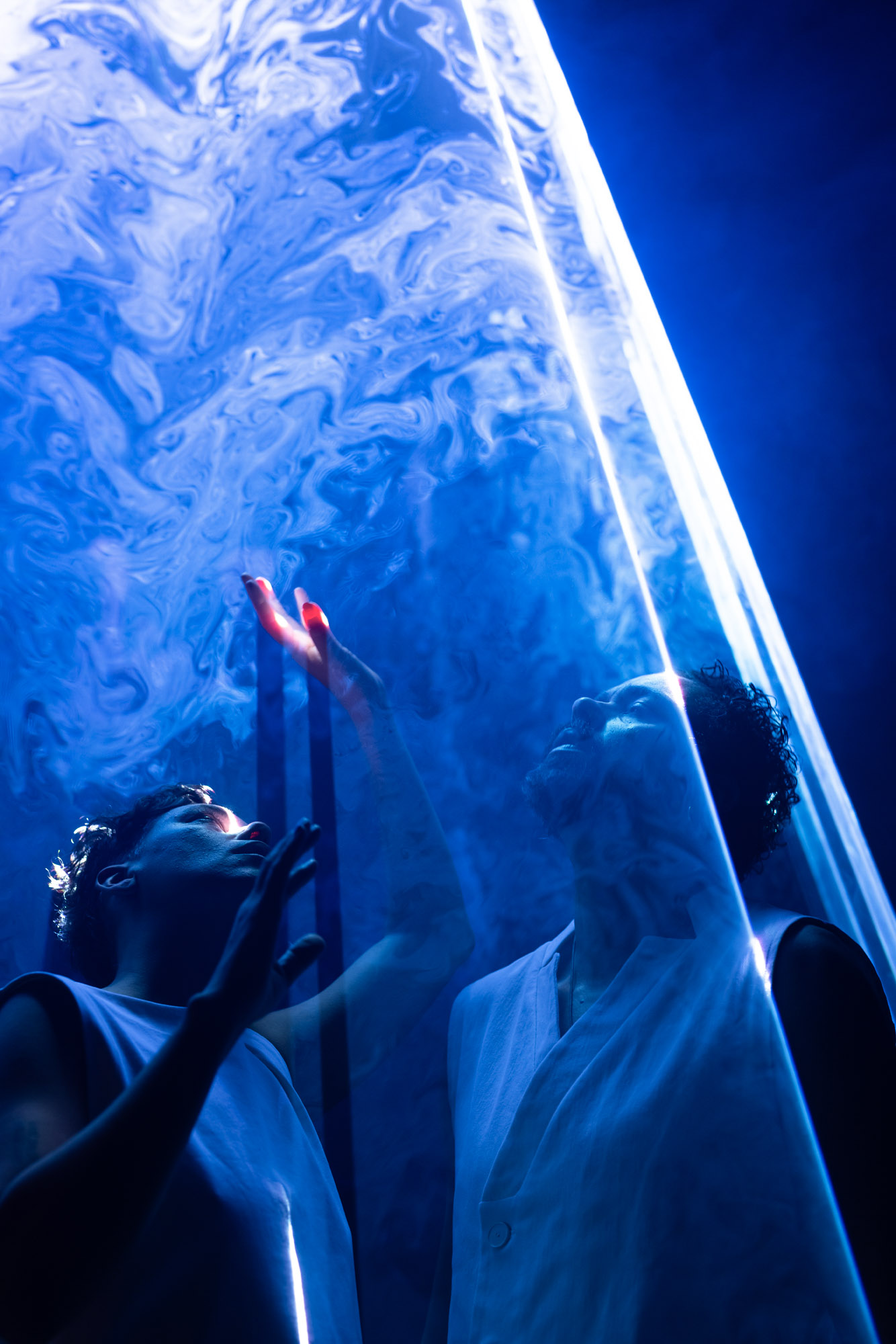 Image description: Two performers stand close to each other, eyes looking upward pensively between beams of blue light, filled with swirling haze. One reaches her fingers into the light, like dipping into another realm.