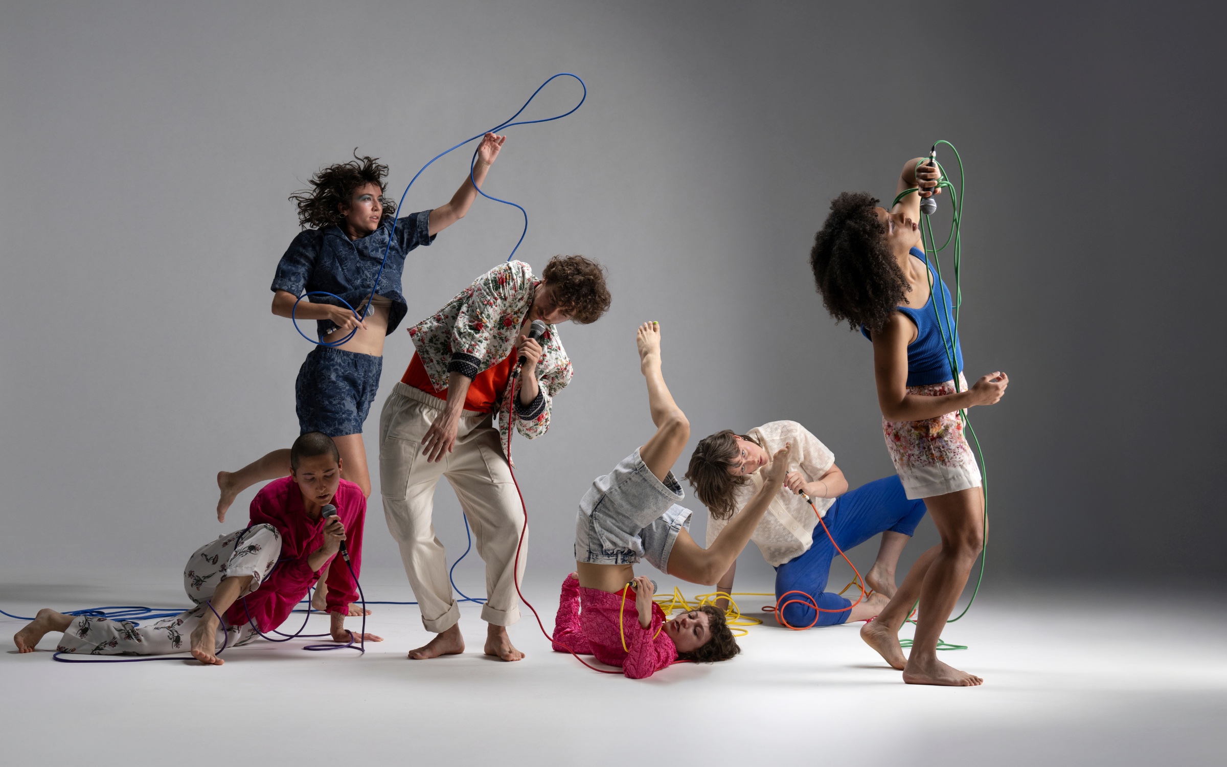 Image description: Six dancers are caught mid-action, each body making a different shape. They hold microphones with long colourful cords that hang and twist between them like squiggles on a page.