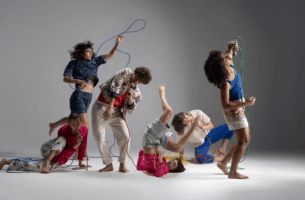 Image description: Six dancers are caught mid-action, each body making a different shape. They hold microphones with long colourful cords that hang and twist between them like squiggles on a page.