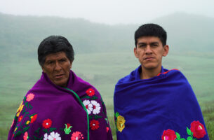 Image description: Outdoors against a backdrop of the misty mountains, two Indigenous men stare forwards with serious, engaging expressions. Both wrap bright blankets, embroidered exquisitely with bold colourful flowers, around their shoulders, covering their bodies and further emphasizing their serious faces.