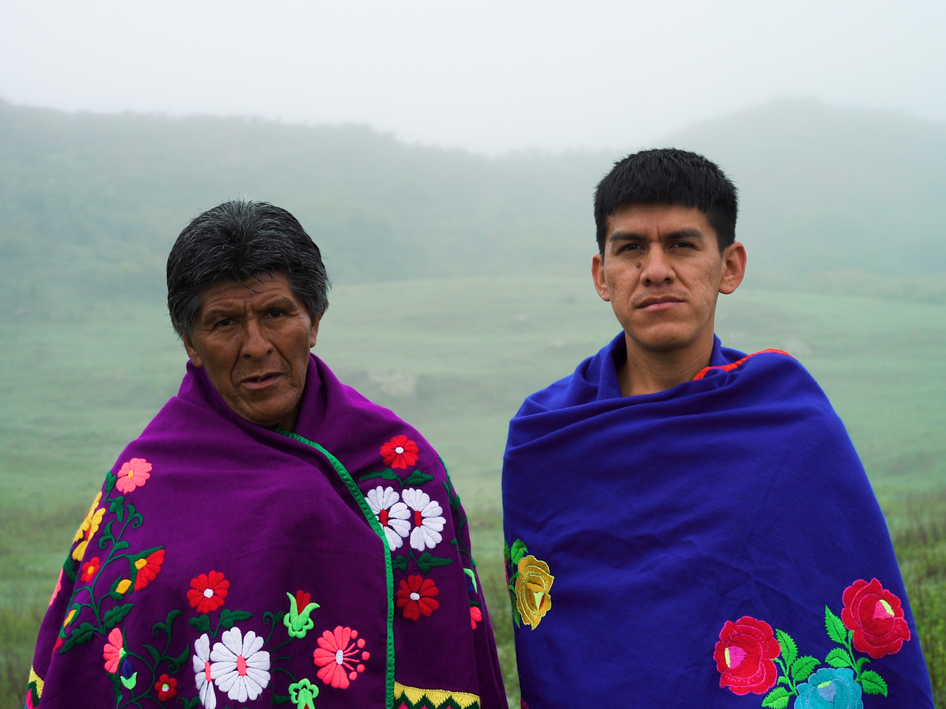 Image description: Outdoors against a backdrop of the misty mountains, two Indigenous men stare forwards with serious, engaging expressions. Both wrap bright blankets, embroidered exquisitely with bold colourful flowers, around their shoulders, covering their bodies and further emphasizing their serious faces.