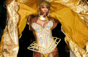 Image description: A Black femme steps onto the runway in gorgeous golden vogue ball attire: impeccable makeup framed by an elegant gold headpice, a structured gold bodice, generous flakes of gold on her tights. She holds up her immense jacket so that a huge swath of luxurious gold fabric drapes over her head and frames her form.