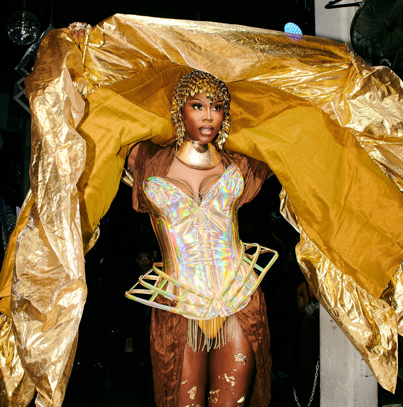 Image description: A Black femme steps onto the runway in gorgeous golden vogue ball attire: impeccable makeup framed by an elegant gold headpice, a structured gold bodice, generous flakes of gold on her tights. She holds up her immense jacket so that a huge swath of luxurious gold fabric drapes over her head and frames her form.