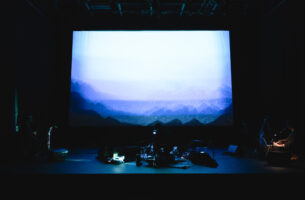 Image description: A performer crouches on a dim stage amidst a complex array of technical equipment, like monitors, projectors and microphone stands. Behind them, an image of a mountainous horizon superimposed over itself creates misty blue layers.