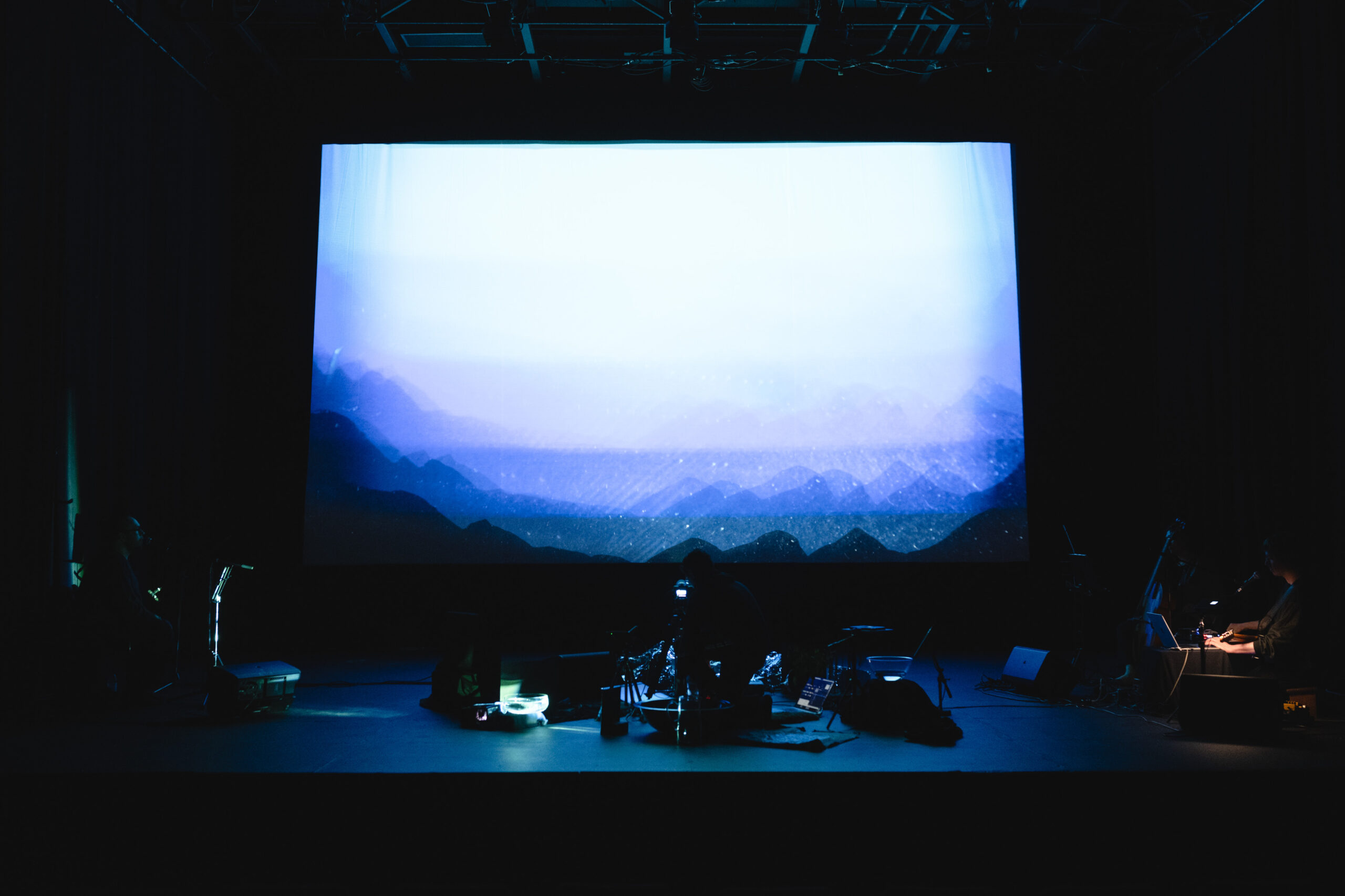 Image description: A performer crouches on a dim stage amidst a complex array of technical equipment, like monitors, projectors and microphone stands. Behind them, an image of a mountainous horizon superimposed over itself creates misty blue layers.