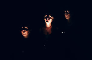 Image description: On a pitch black stage, three women's faces are illuminated from below with flashlights. Their shadowy expressions are serious, as though peering into an unknown darkness.
