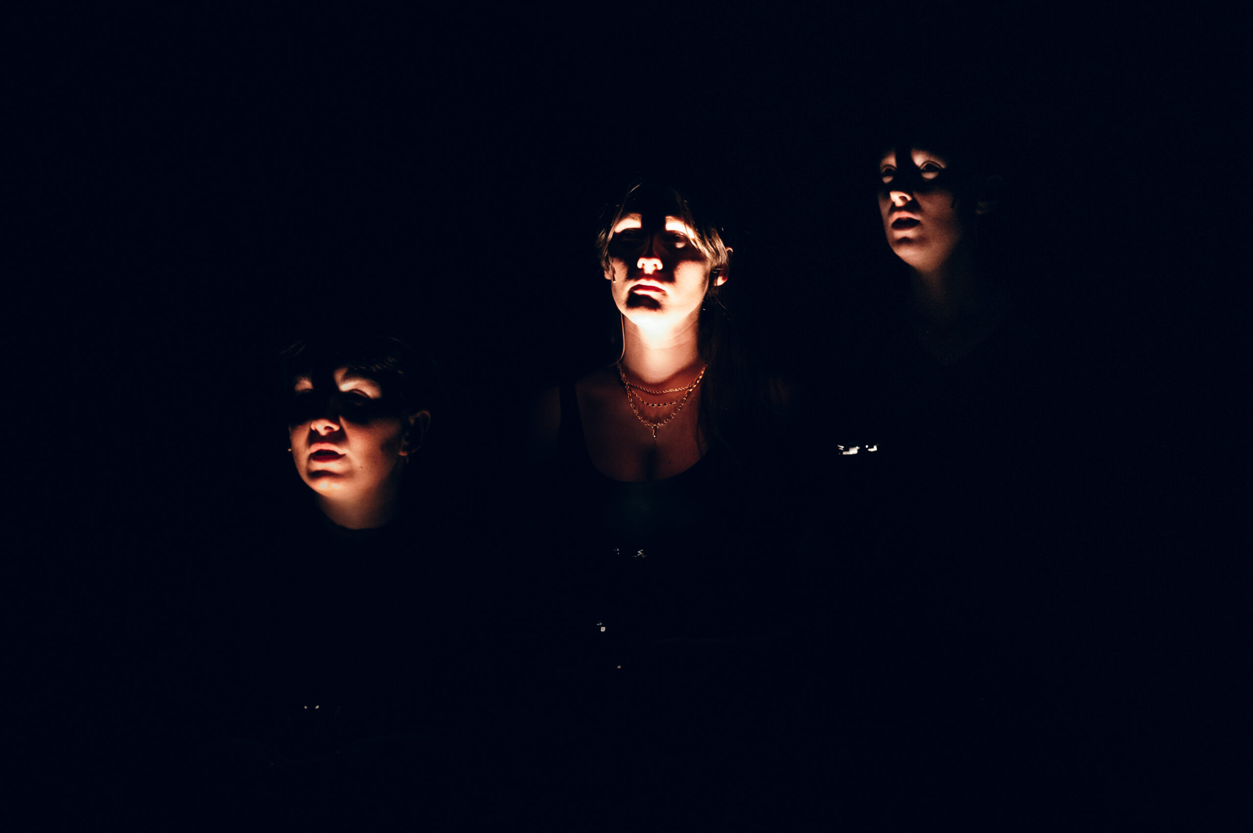 Image description: On a pitch black stage, three women's faces are illuminated from below with flashlights. Their shadowy expressions are serious, as though peering into an unknown darkness.