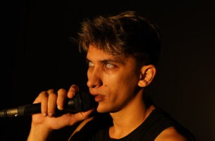 Image description: A young man stares out from stage with intensity holding a microphone. His eyes have no colour, giving him the appearance of being haunted. Warm light bathes his olive skin.