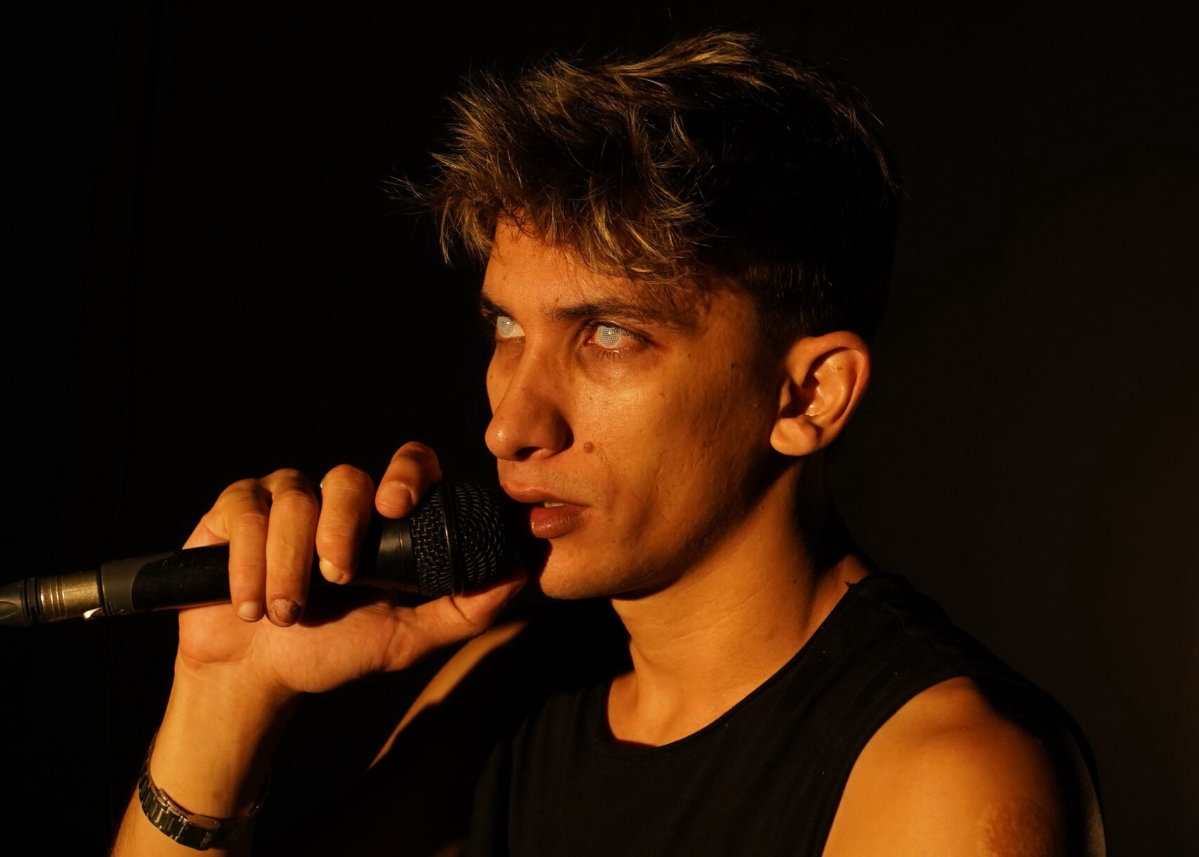 Image description: A young man stares out from stage with intensity holding a microphone. His eyes have no colour, giving him the appearance of being haunted. Warm light bathes his olive skin.