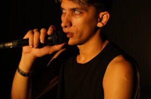 Image description: A young man stares out from stage with intensity holding a microphone. His eyes have no colour, giving him the appearance of being haunted. Warm light bathes his olive skin.