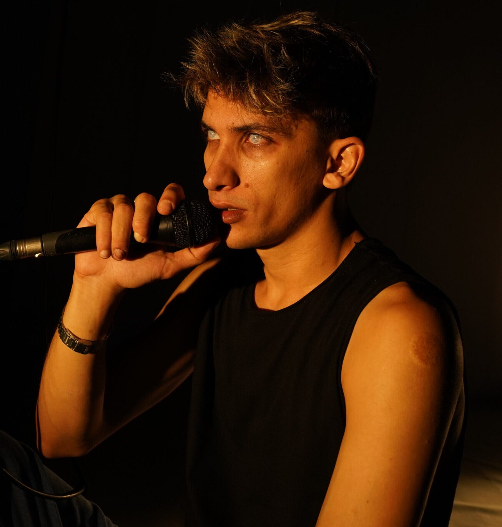 Image description: A young man stares out from stage with intensity holding a microphone. His eyes have no colour, giving him the appearance of being haunted. Warm light bathes his olive skin.