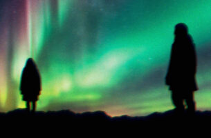 Image description: Two figures are silouetted on a ridge against the bright background of the northern lights, which fills the sky with glowing greens and pinks.